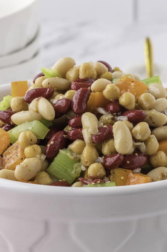 A white bowl filled with a bean salad containing kidney beans, white beans, celery, and diced peppers sits on a marble counter in a kitchen setting.