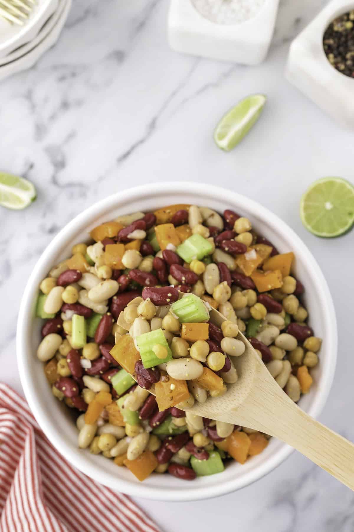 A wooden spoon holds a portion of bean salad with celery and bell pepper over a white bowl filled with the same salad, on a marble countertop with lime wedges nearby.