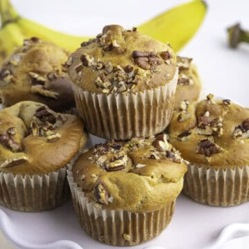 Five banana protein muffins with chopped pecans on top are arranged on a white plate, with bananas visible in the background.