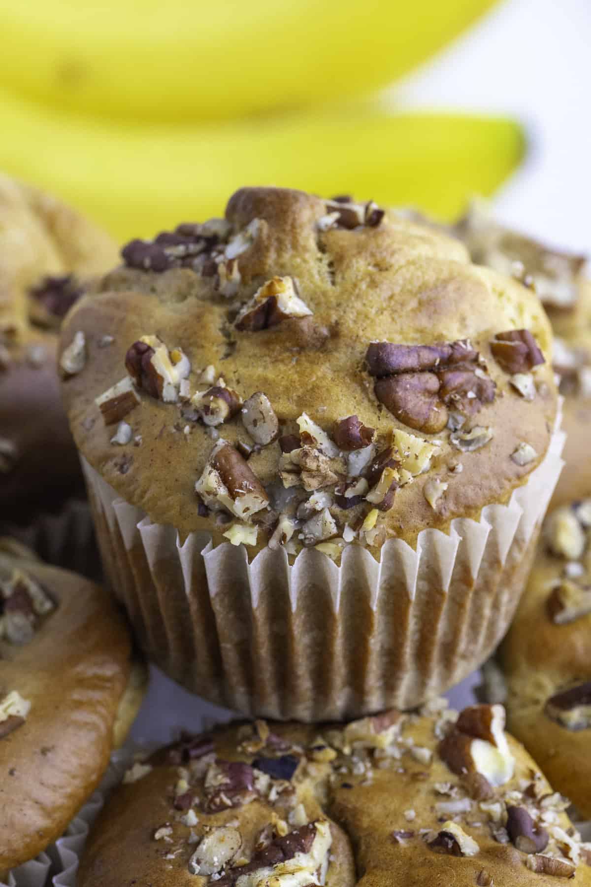 A close-up of a banana protein muffin topped with chopped nuts, surrounded by other muffins, with bananas in the background.