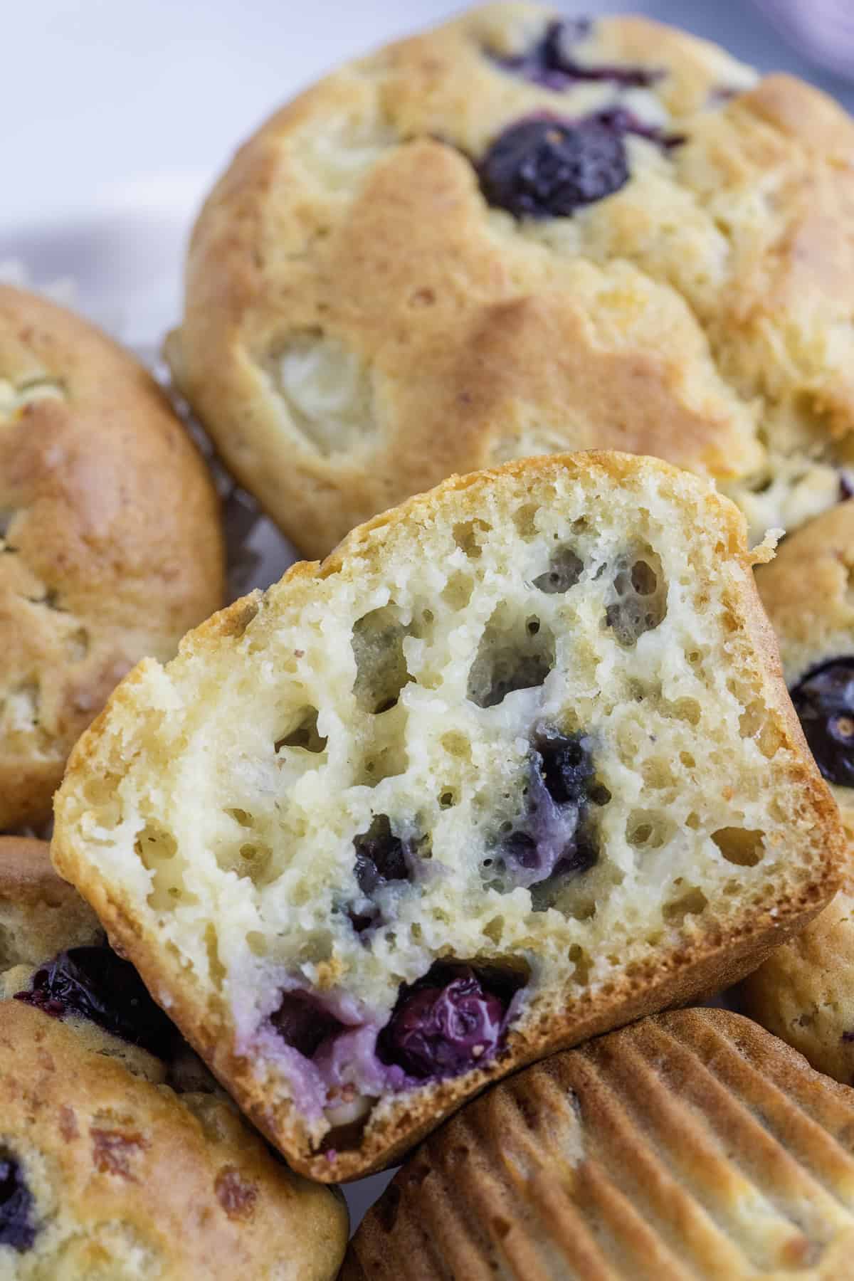 A close-up of blueberry muffins, one of which is cut in half to show the moist, airy interior with visible blueberries.
