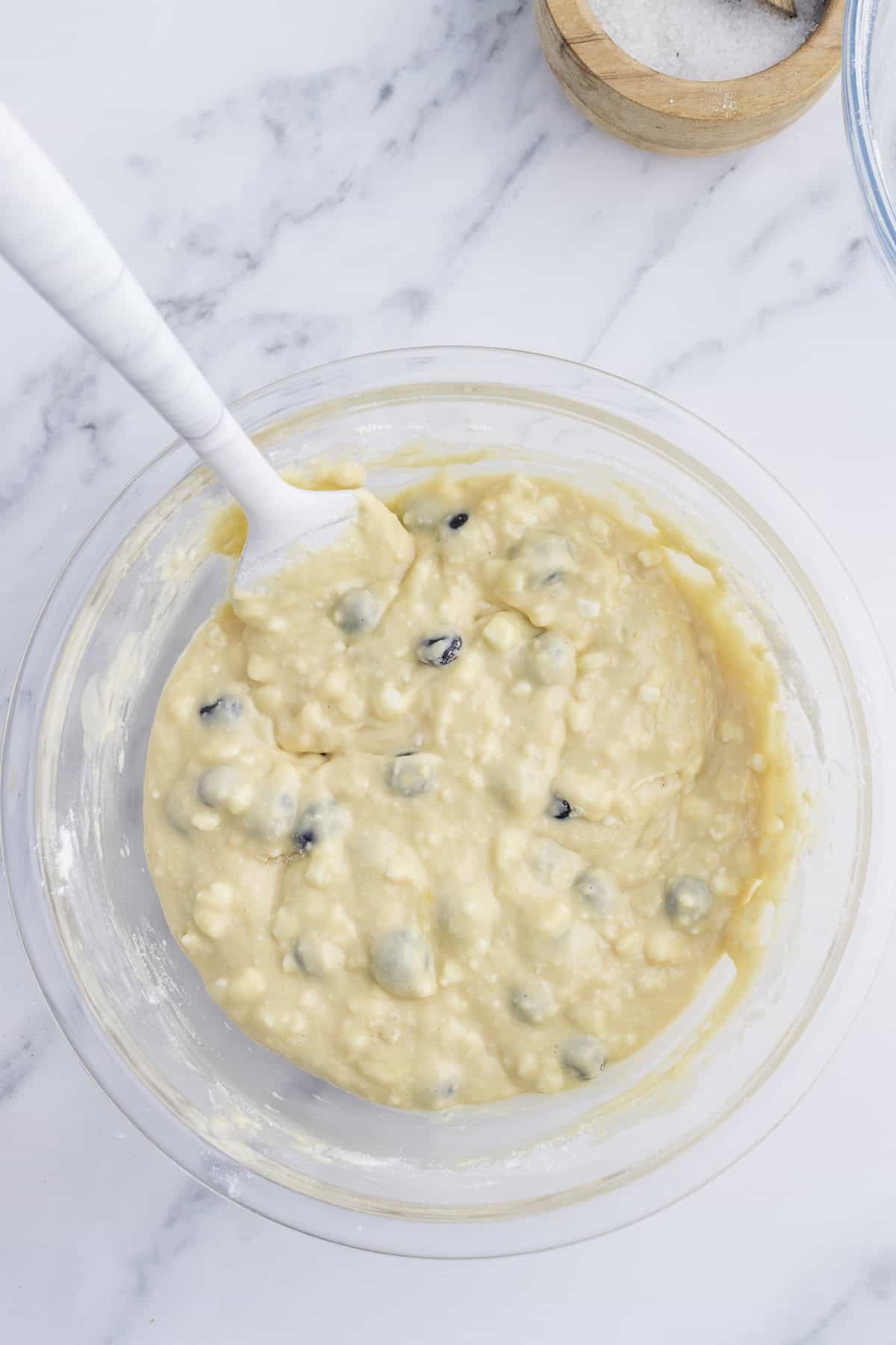 A glass bowl with blueberry muffin batter being mixed with a white spatula on a marble countertop.