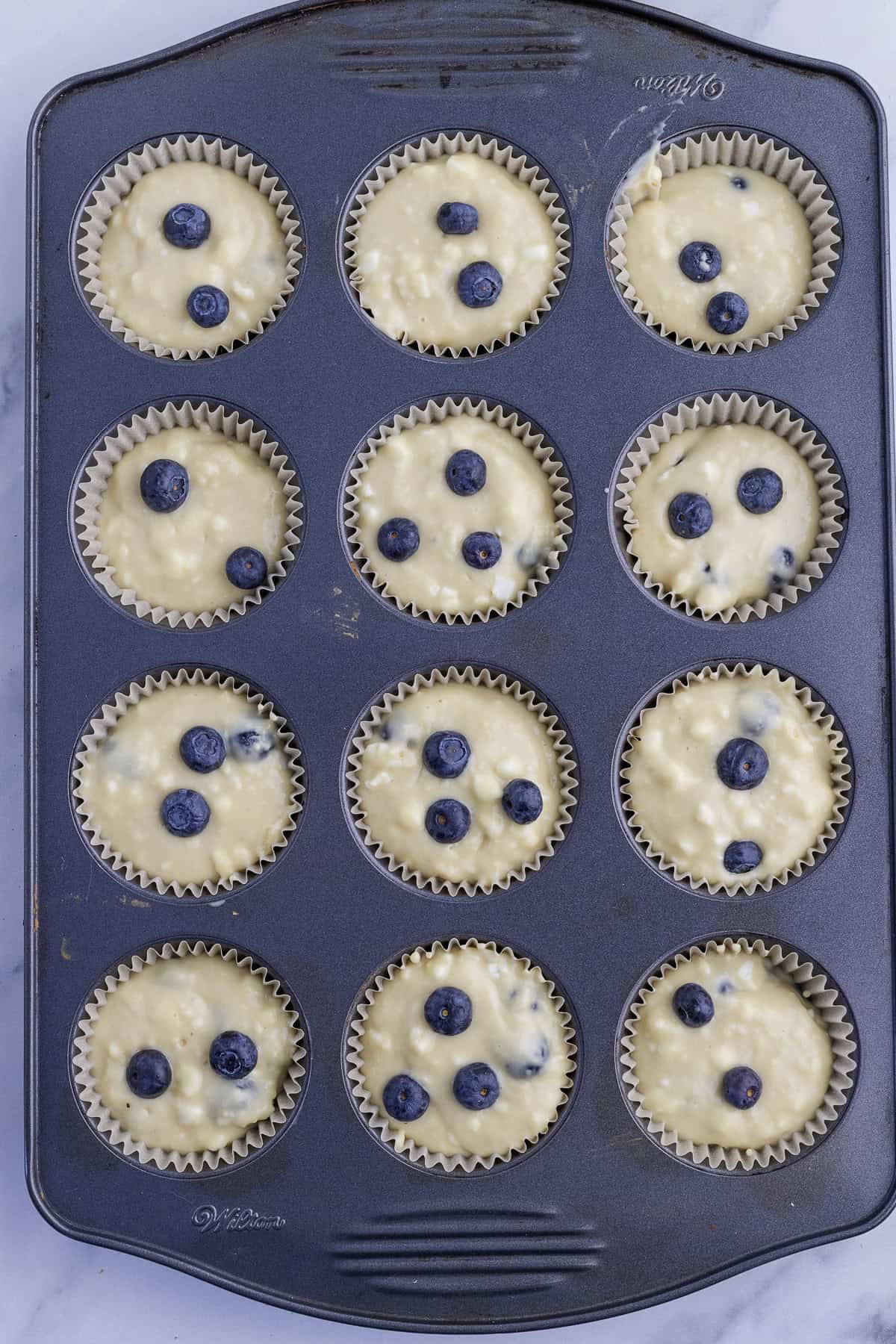 A muffin tin with twelve paper liners filled with blueberry muffin batter, each topped with several whole blueberries, ready to be baked.