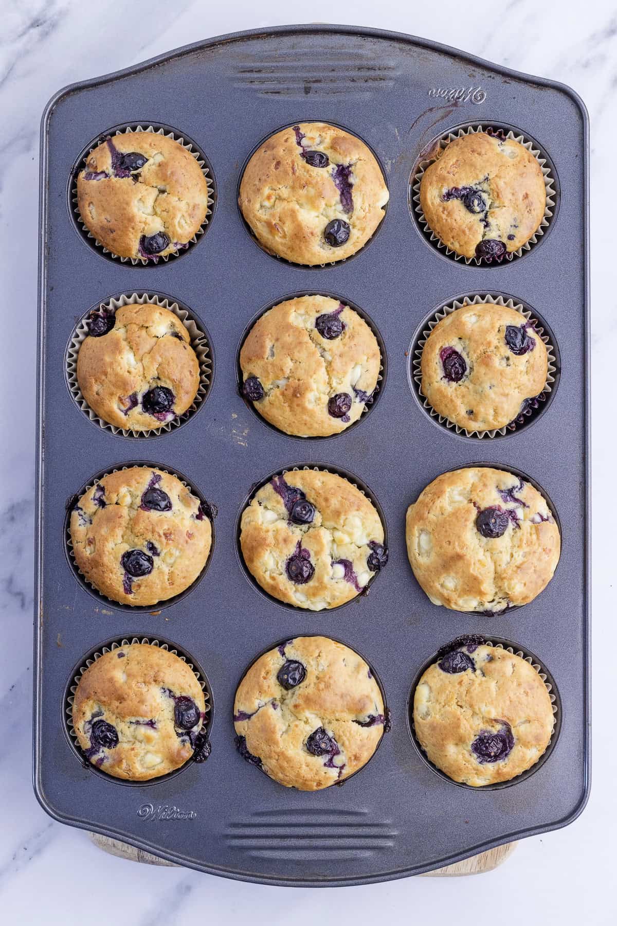 A muffin tin containing twelve freshly baked blueberry muffins, some with browned tops and visible blueberries, sits on a marble surface.
