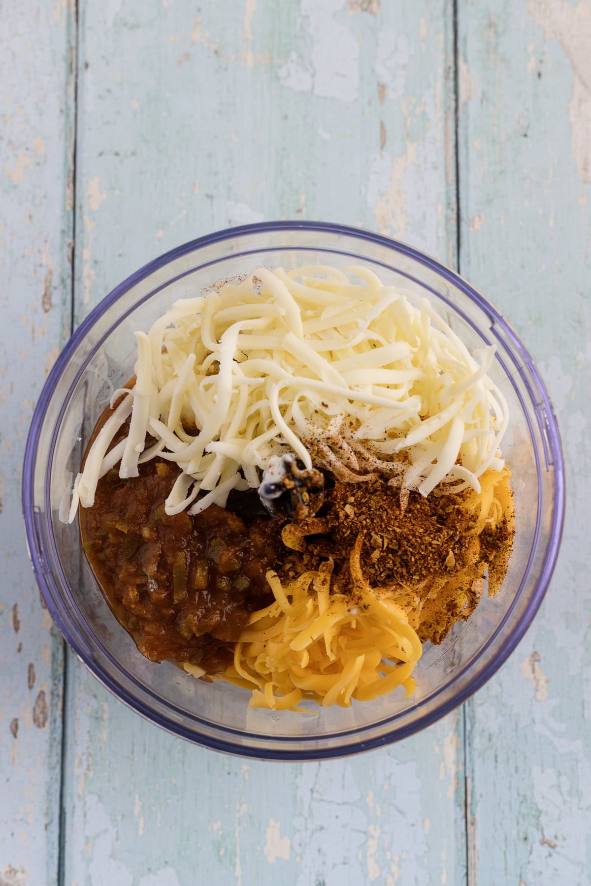 A mixing bowl contains Cottage Cheese, salsa, and  spices on a light blue wooden surface.