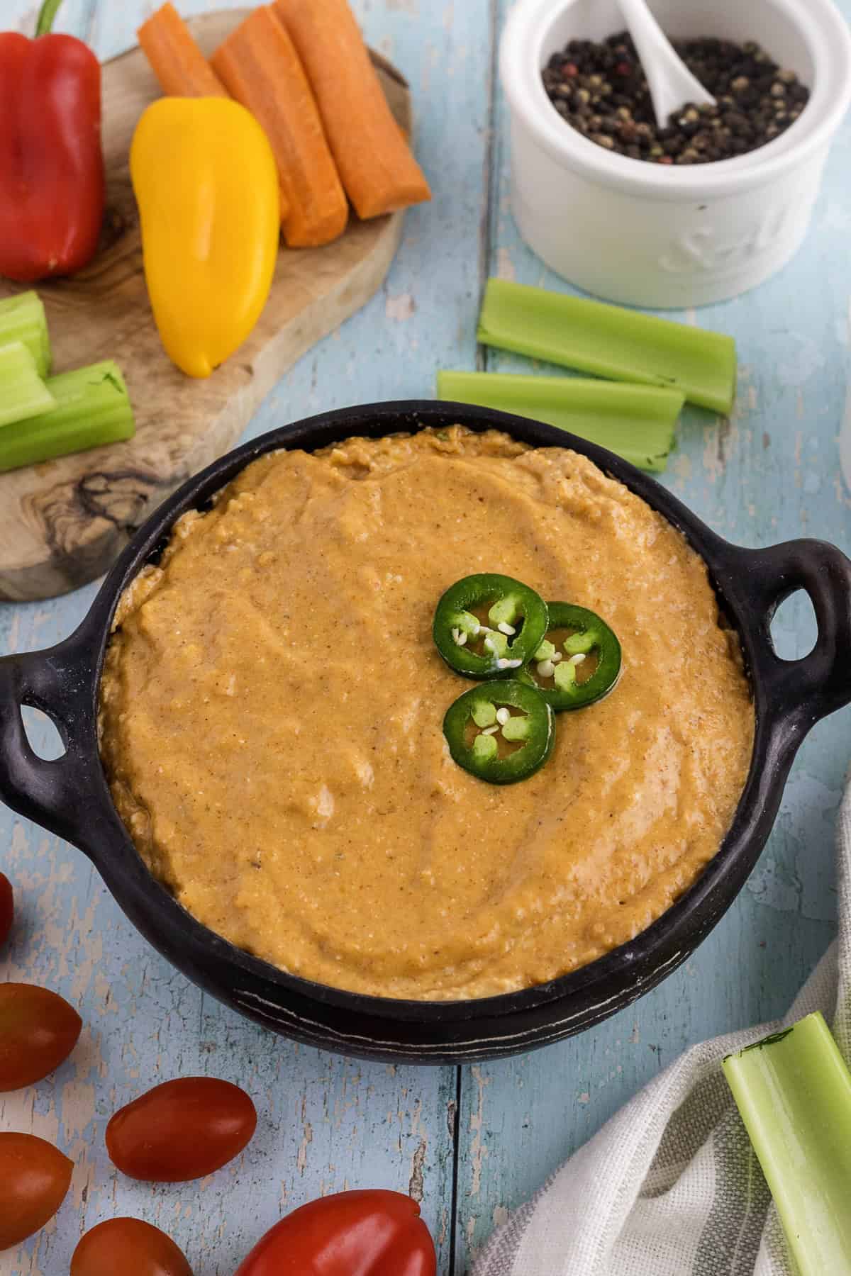 A bowl of creamy Queso Dip topped with sliced jalapeños, surrounded by celery sticks, cherry tomatoes, carrots, bell peppers, and a small bowl of peppercorns.