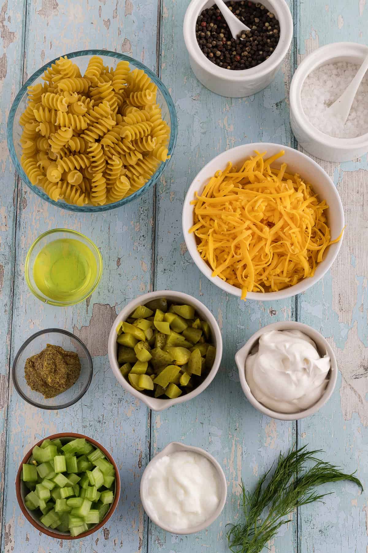 A flat lay of ingredients on a blue wooden surface, including rotini pasta, shredded cheddar, celery, pickles, spices, oil, sour cream, and dill.