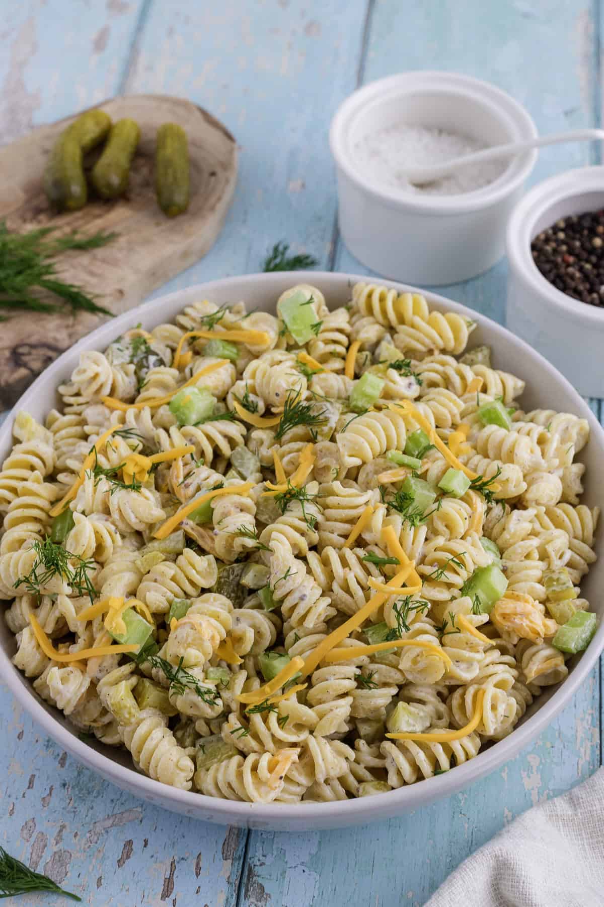 A bowl of creamy rotini pasta salad with shredded cheese, chopped pickles, and fresh dill, placed on a blue wooden surface with salt, pepper, and pickles on the side.