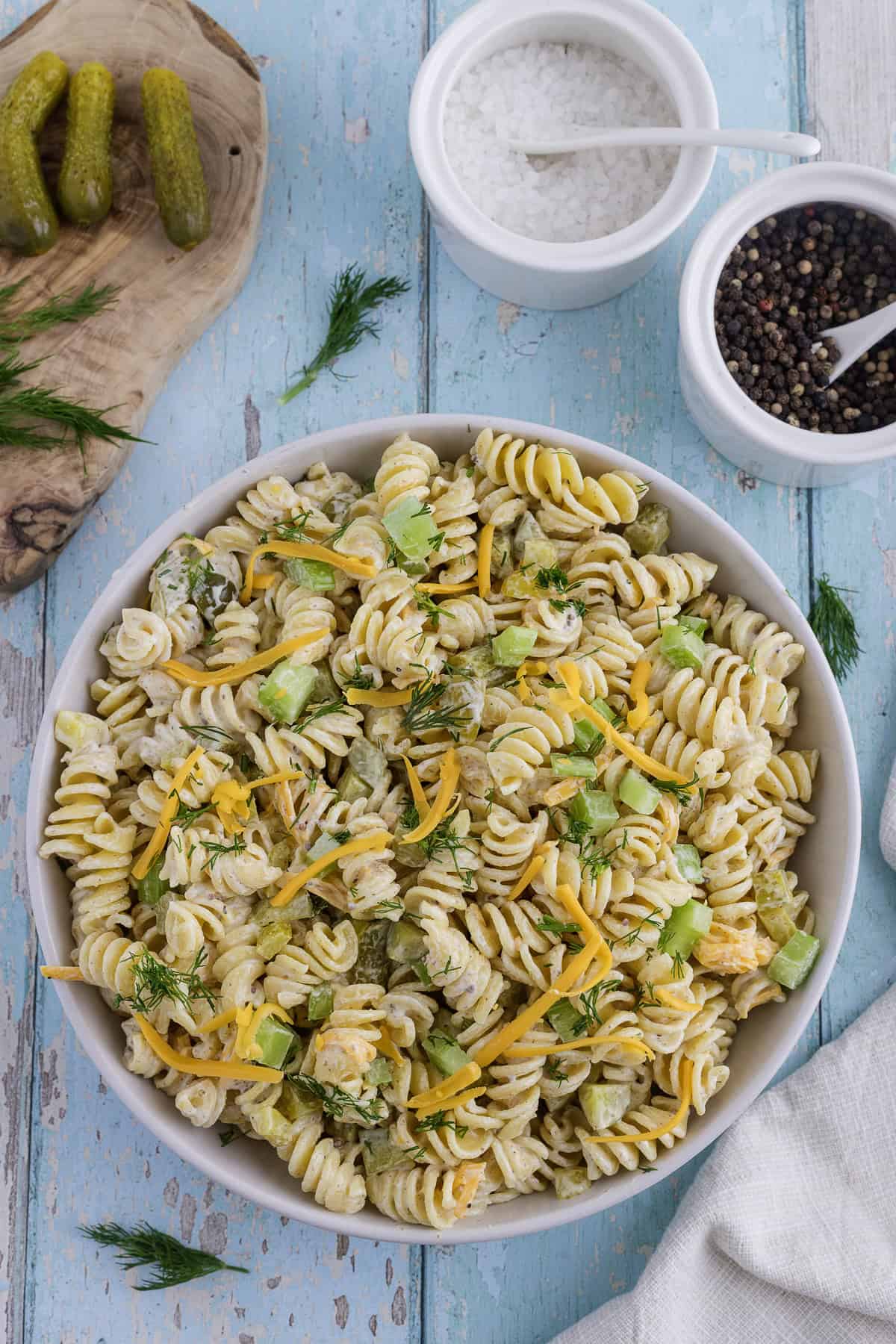A bowl of rotini pasta salad with shredded cheese, chopped pickles, celery, and dill, next to small bowls of salt, pepper, and whole pickles on a rustic blue wooden surface.