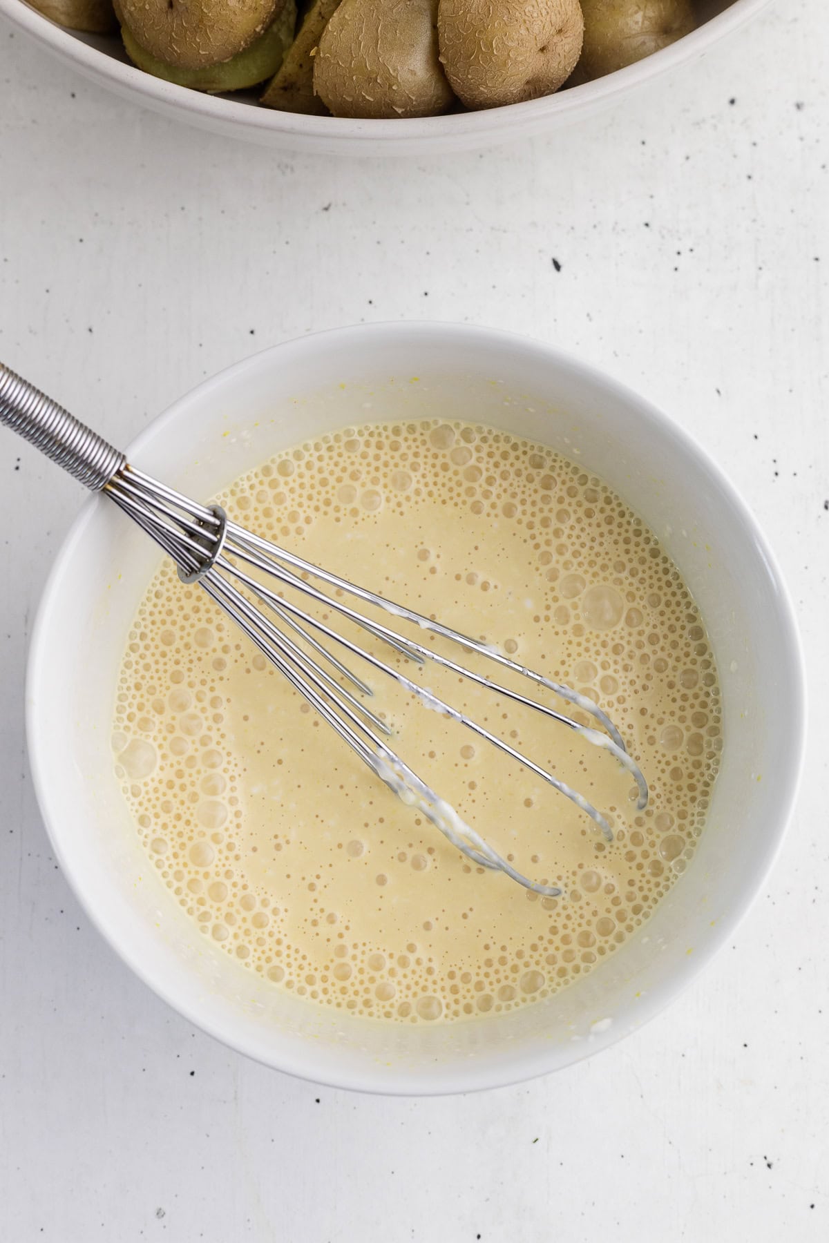 A metal whisk resting in a white bowl containing a pale yellow, frothy liquid mixture sits on a white surface.