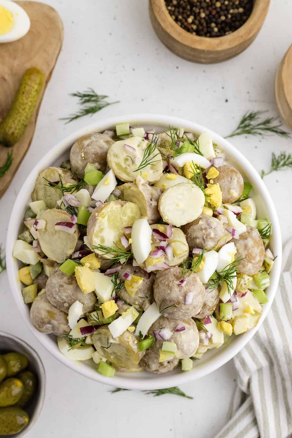A bowl of potato salad with sliced hard-boiled eggs, chopped red onion, celery, and fresh dill, surrounded by bowls of spices and pickles on a white surface.
