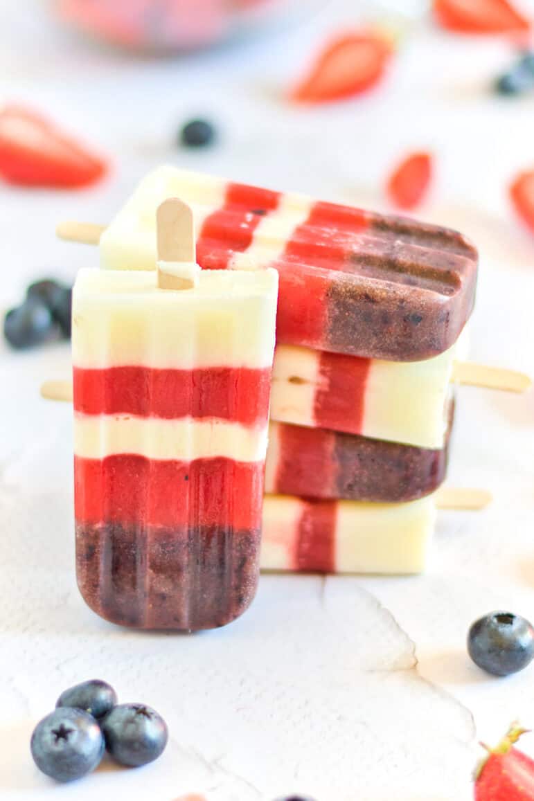 Three striped popsicles with layers of white, red, and purple are stacked on a white surface, surrounded by scattered blueberries and strawberry pieces.