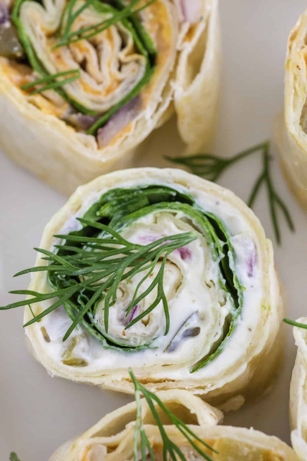 A close-up of a tortilla pinwheel filled with spinach, cream cheese, and herbs, garnished with fresh dill.