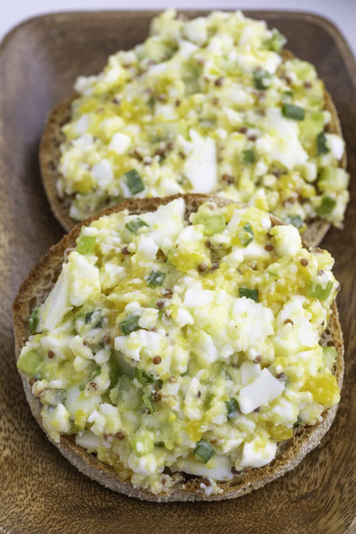 Two halves of a whole grain English muffin topped with egg salad containing chopped eggs, green onions, and mustard, served on a wooden plate.