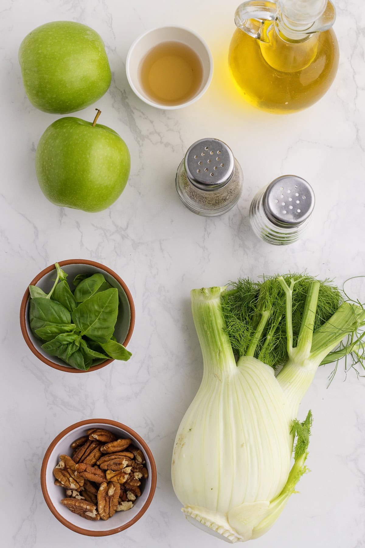 Top-down view of fennel bulb, green apples, basil leaves, pecans, salt, pepper, oil, and vinegar arranged on a white marble surface.