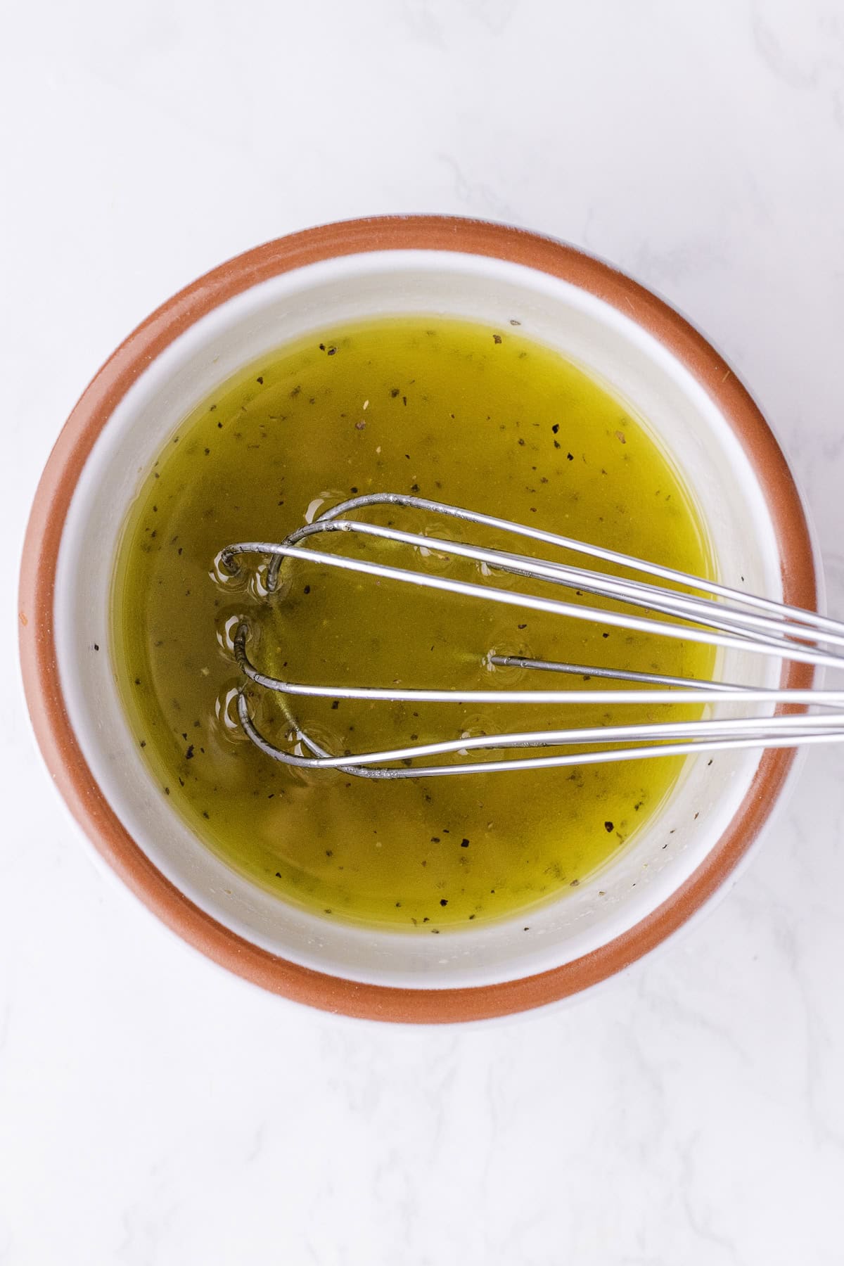 Un fouet en métal posé dans un bol de vinaigrette jaune-vert aux herbes, posé sur une surface en marbre blanc.
