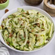 Bowl of salad with sliced green apples, fennel, herbs, and chopped pecans, seasoned with pepper and served on a white surface.