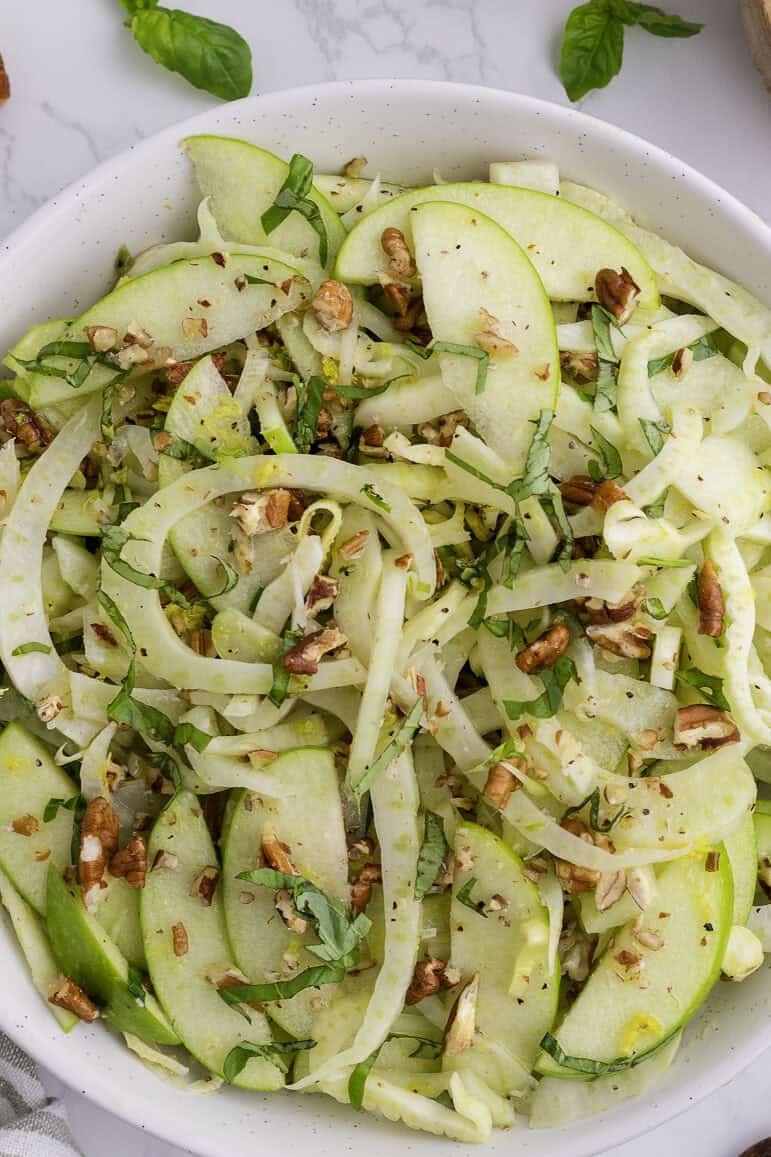 Une salade avec des pommes vertes finement tranchées, du fenouil, des herbes fraîches et des pacanes hachées dans un bol blanc sur une surface en marbre.