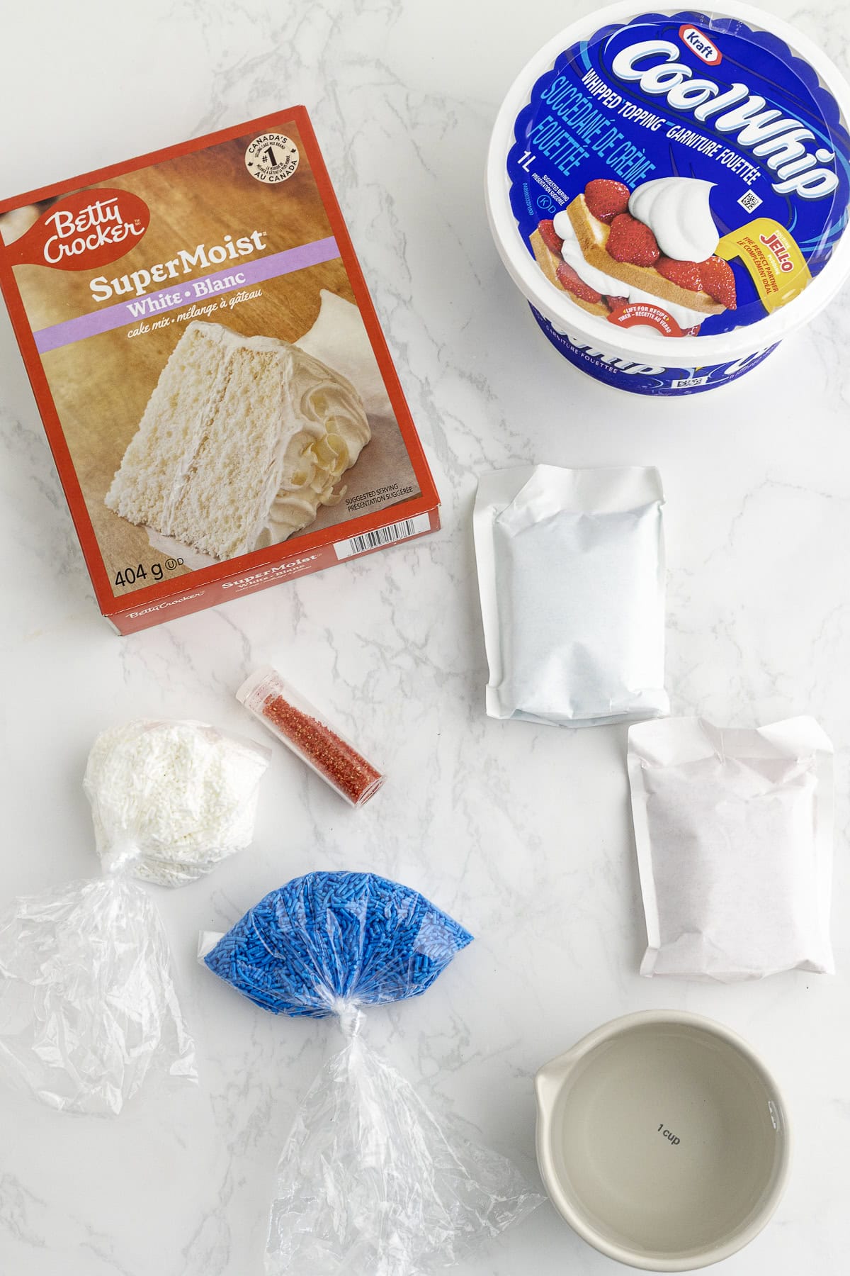 Ingredients for a cake including white cake mix, whipped topping, sprinkles,jello, and water on a marble surface.