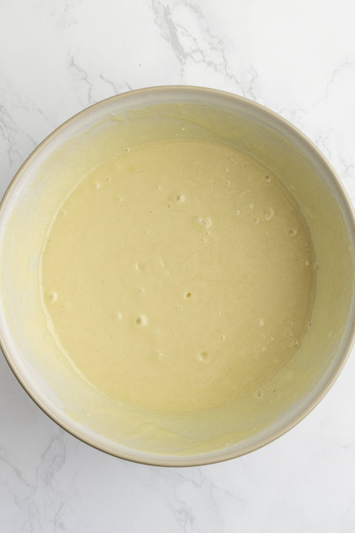 A mixing bowl filled with smooth, pale yellow batter on a white marble surface.