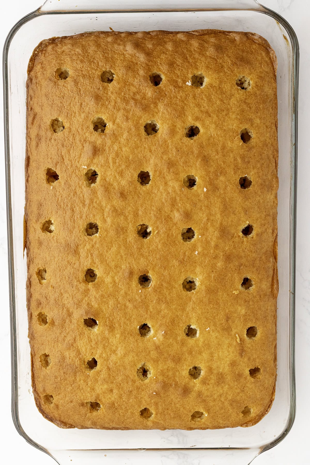 A rectangular golden-brown cake in a glass baking dish with evenly spaced holes poked across the surface.