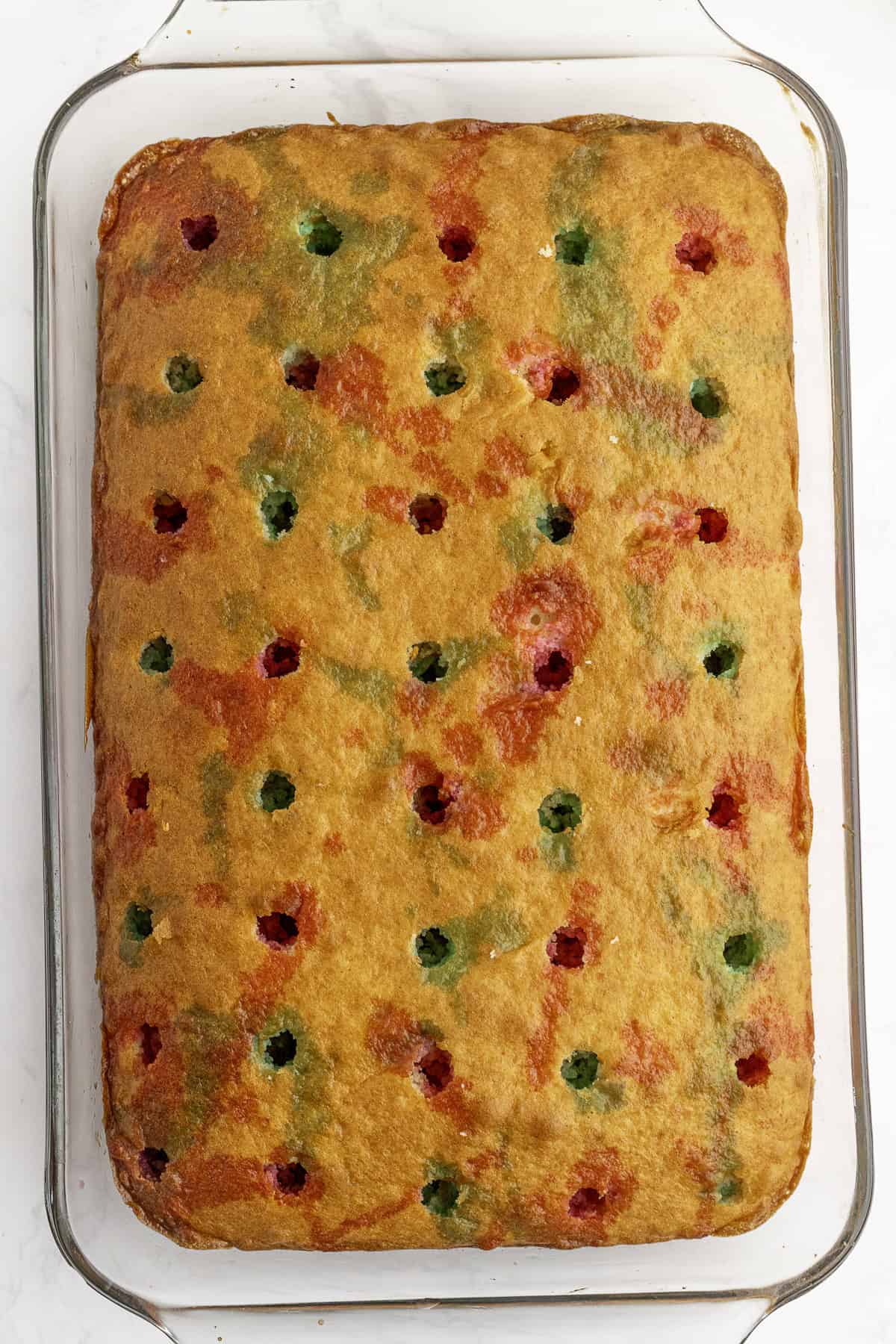 Rectangular baked cake in a glass dish with evenly spaced holes on the surface, some filled with red and green coloring.