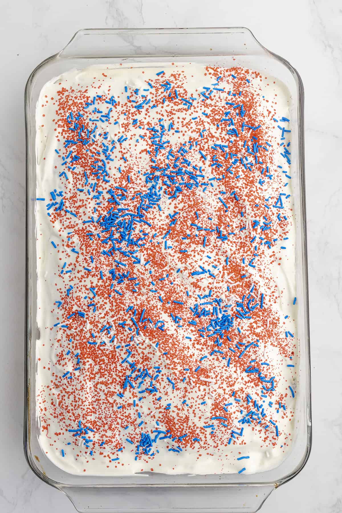 A rectangular glass dish with white frosted cake, topped with red and blue sprinkles, on a white marble surface.