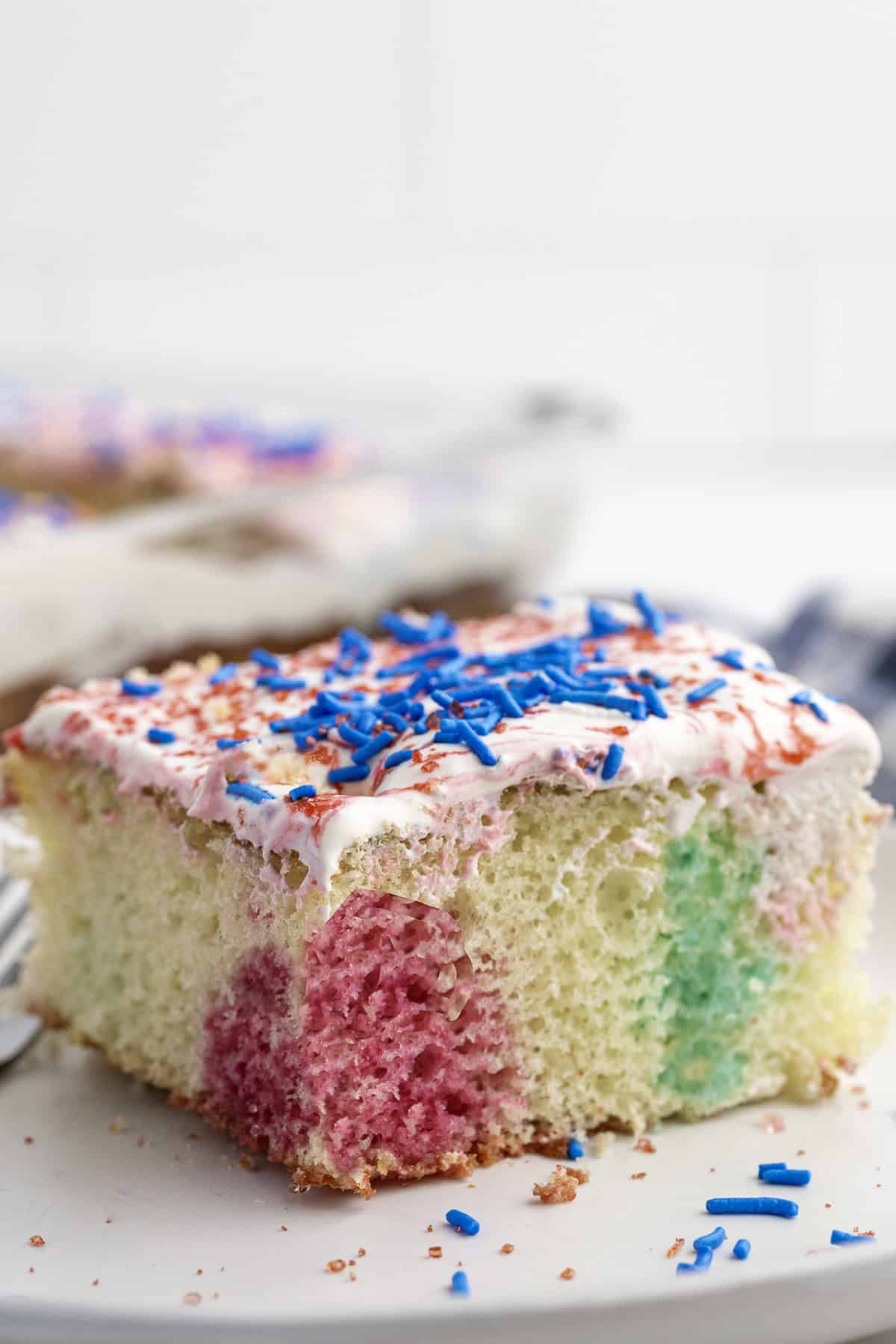 A slice of cake with white icing, blue sprinkles, and colorful patches of pink and green in the sponge on a white plate.