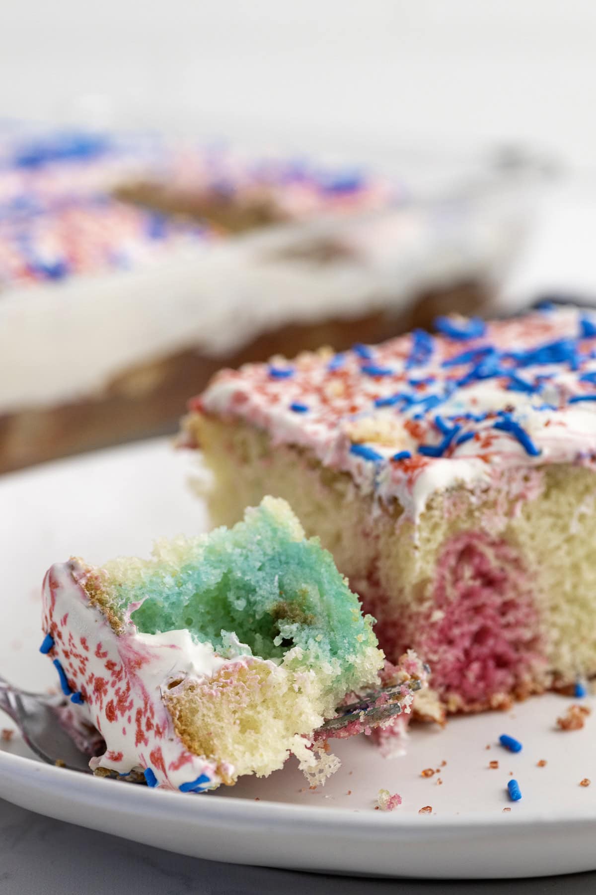 A slice of vanilla cake with red, white, and blue sprinkles and swirls, topped with white frosting, sits on a plate with a bite on a fork.