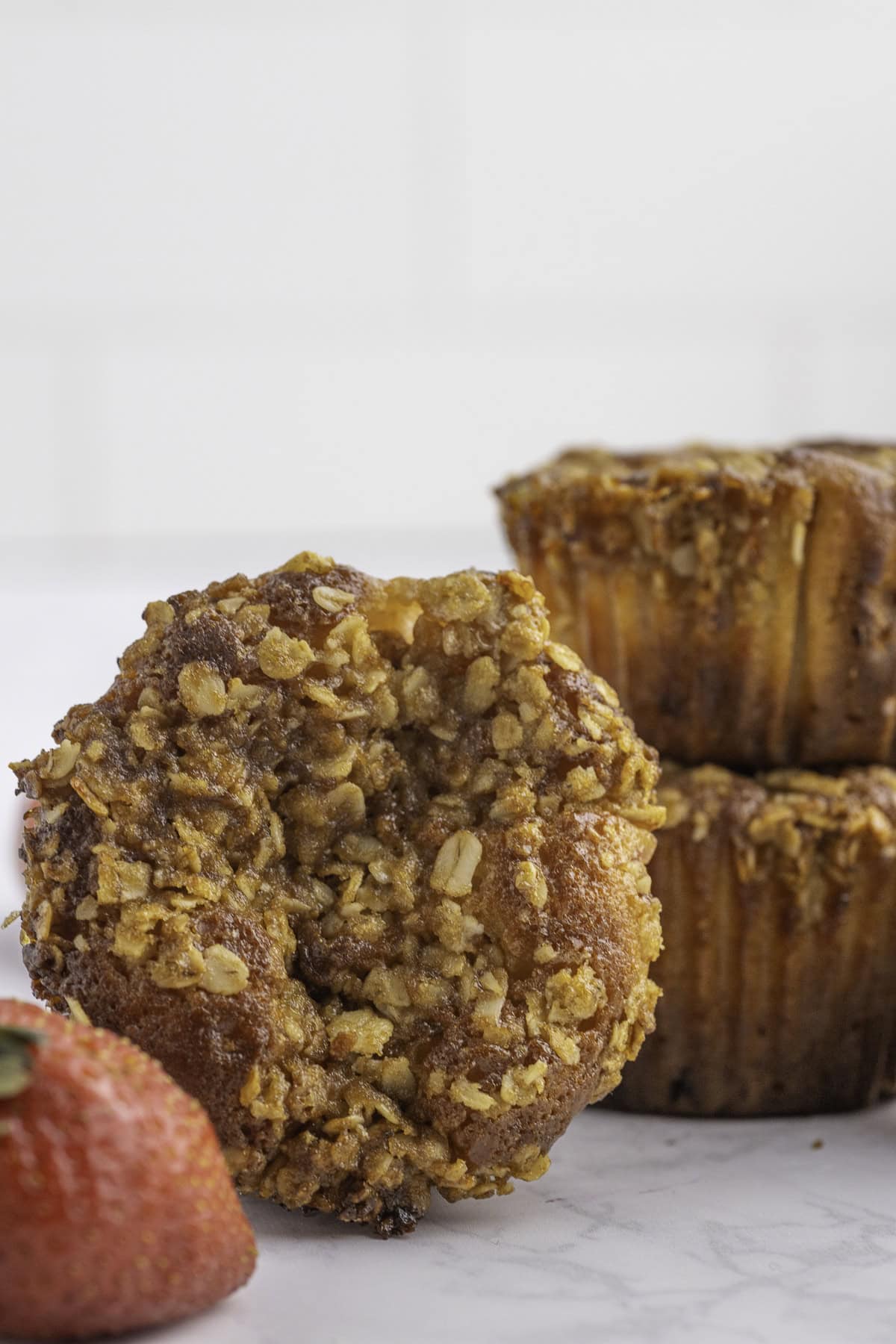 A close-up of an oatmeal-topped muffin is shown in front of a stack of similar muffins, with part of a strawberry visible in the foreground.