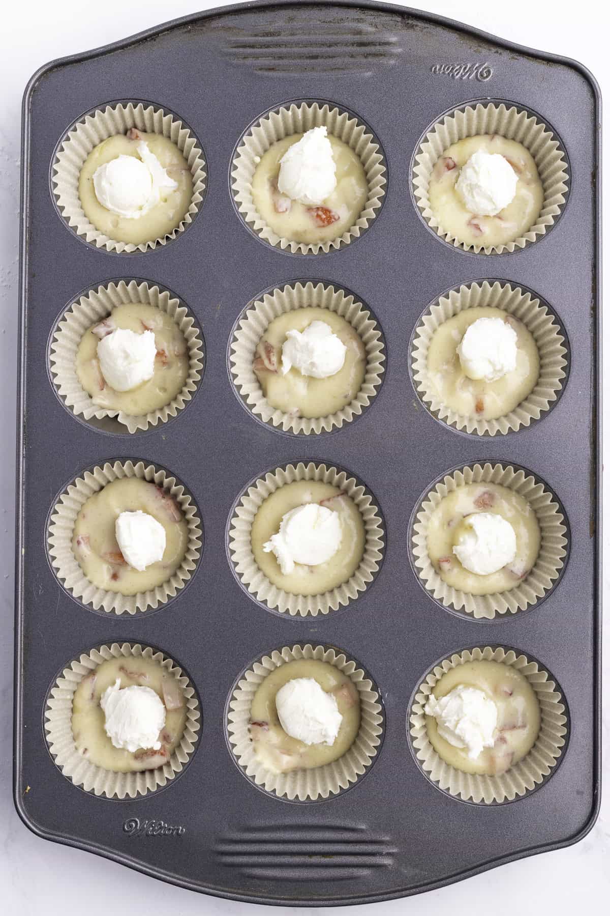 A muffin tin with twelve paper liners, each filled with batter and topped with a dollop of white cream cheese mixture, ready to bake.