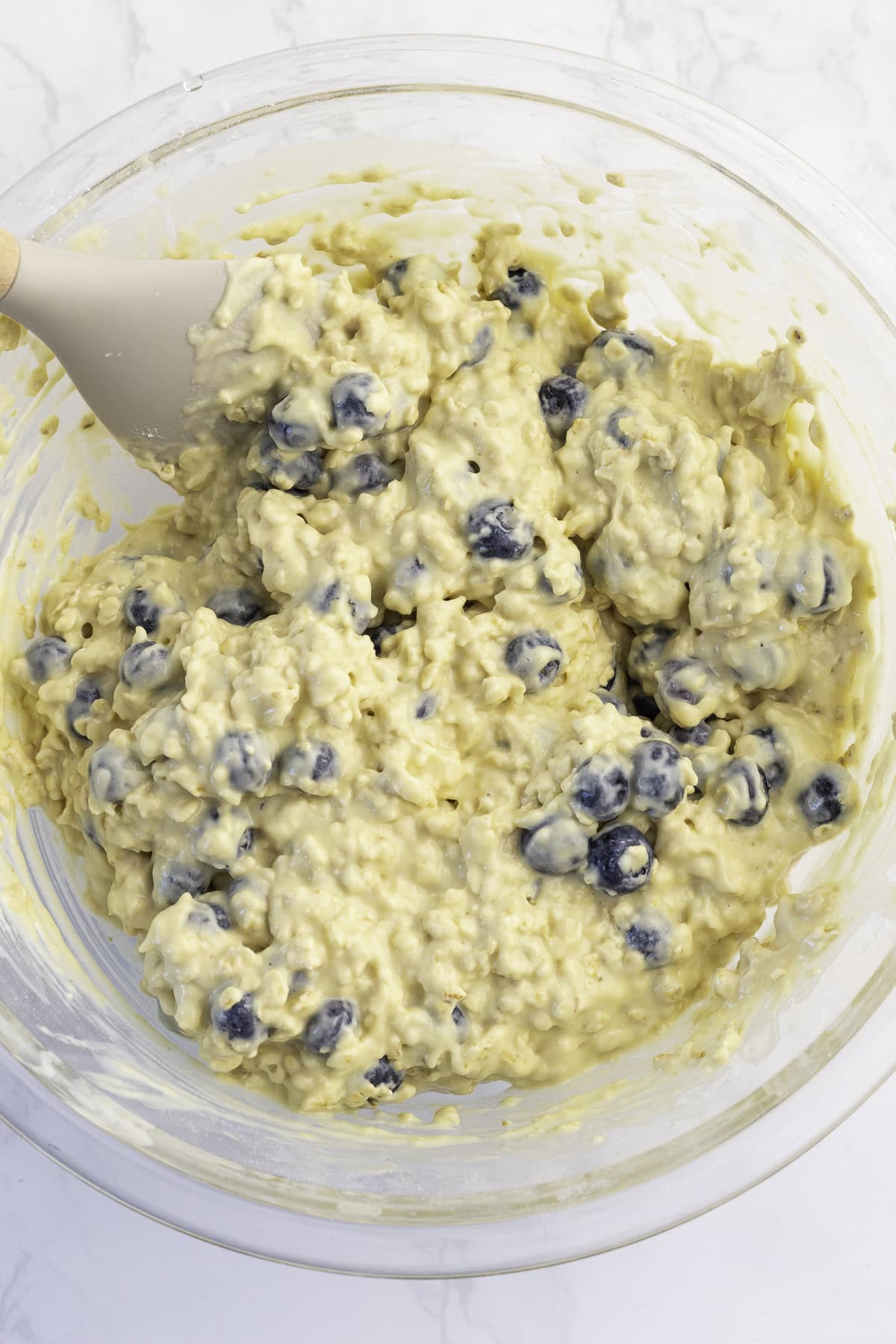 A glass bowl filled with thick batter containing blueberries is being mixed with a spatula.