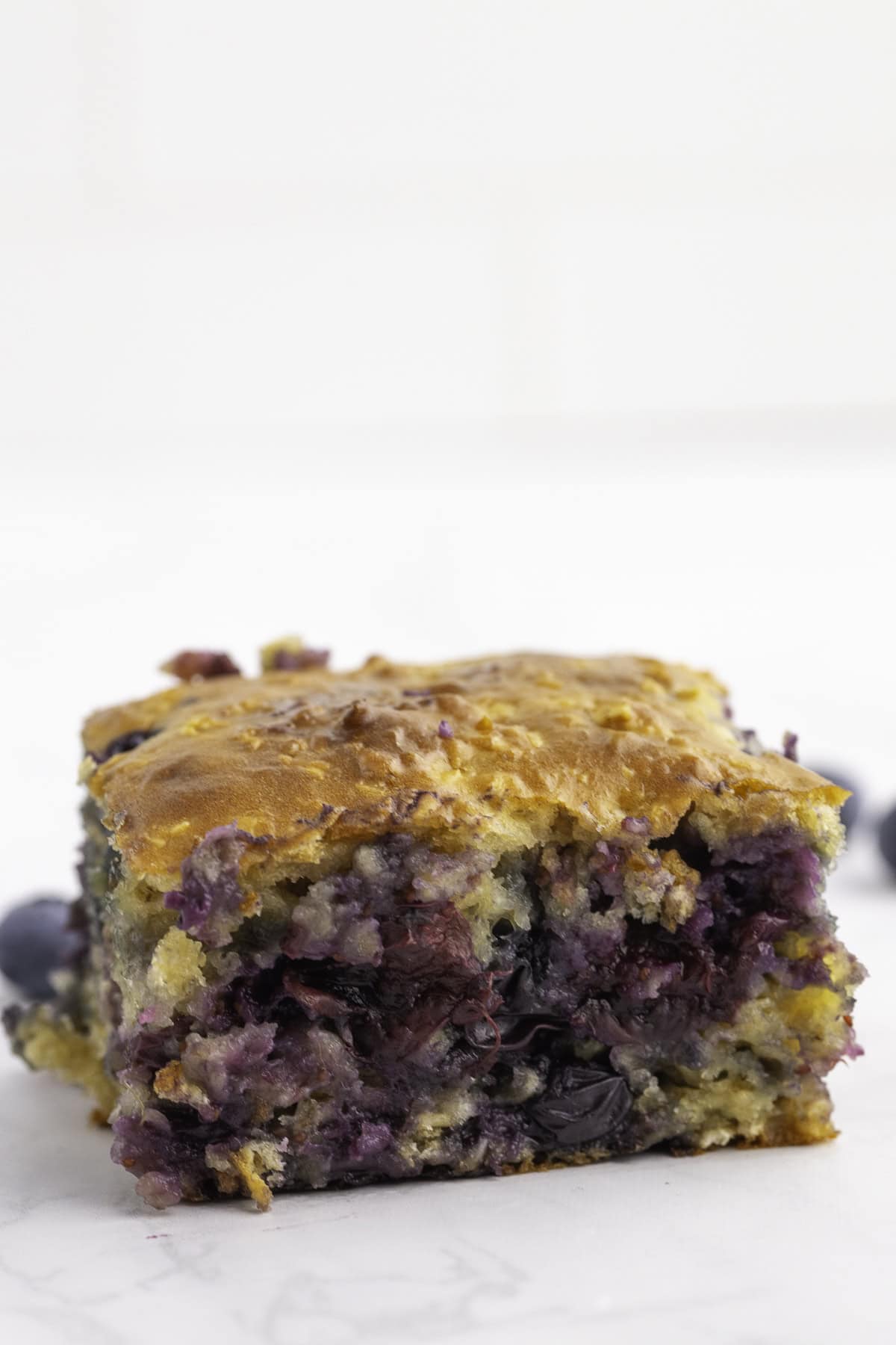 A close-up of a single square piece of blueberry bread with a golden-brown top and visible blueberries inside, placed on a white surface.