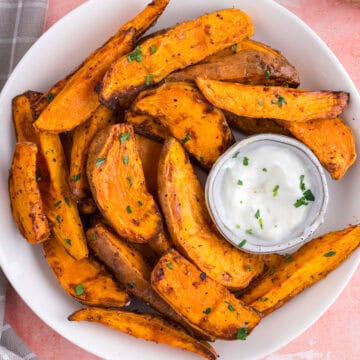 A white plate of seasoned sweet potato wedges garnished with herbs, served with a small dish of white dipping sauce.