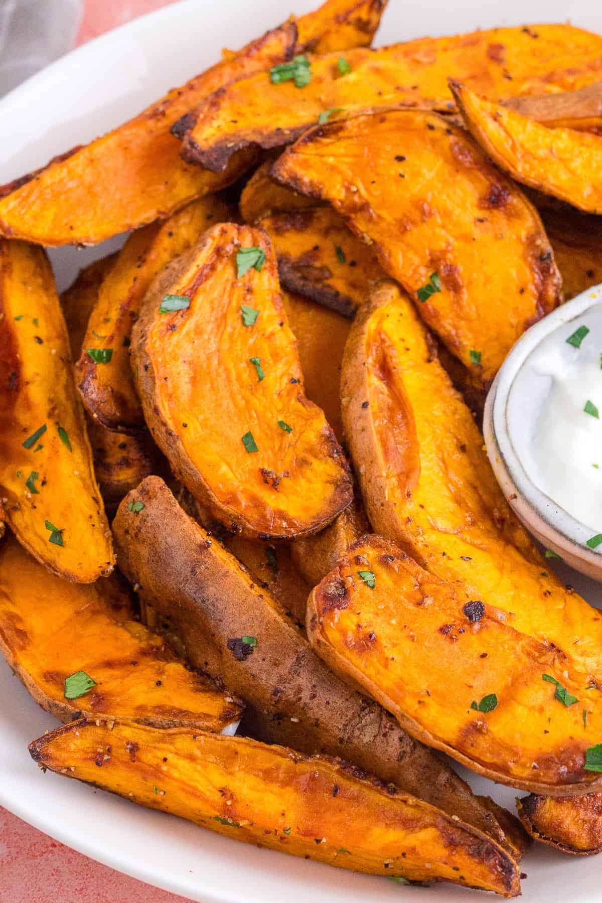 A plate of baked sweet potato wedges garnished with chopped herbs, served with a side of white dipping sauce.