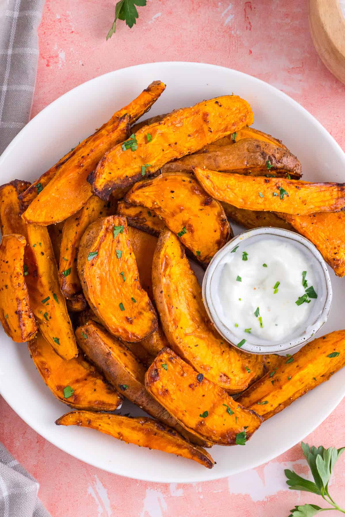 A white plate with baked sweet potato wedges garnished with herbs, served with a small container of white dipping sauce.