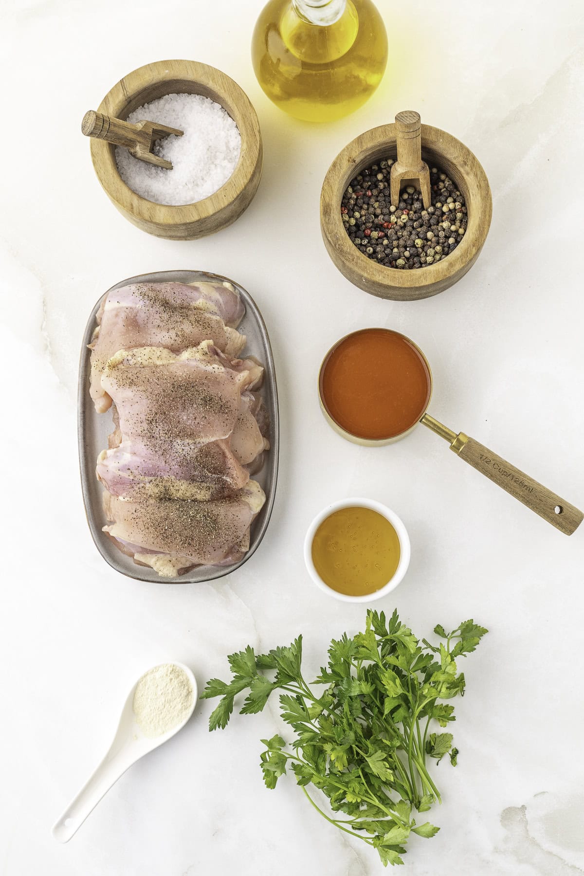 Raw chicken thighs on a tray, surrounded by bowls of salt, pepper, olive oil, buffalo sauce, honey, garlic powder, and fresh parsley on a white surface.