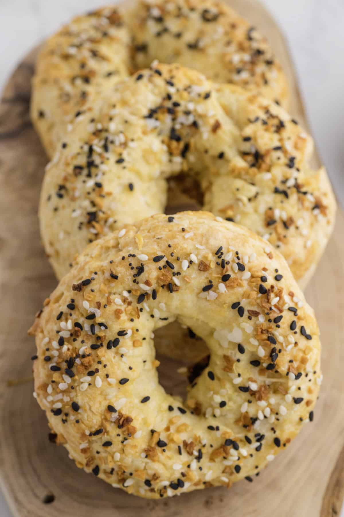 Three everything bagels with a golden crust and assorted seeds sit on a wooden board.