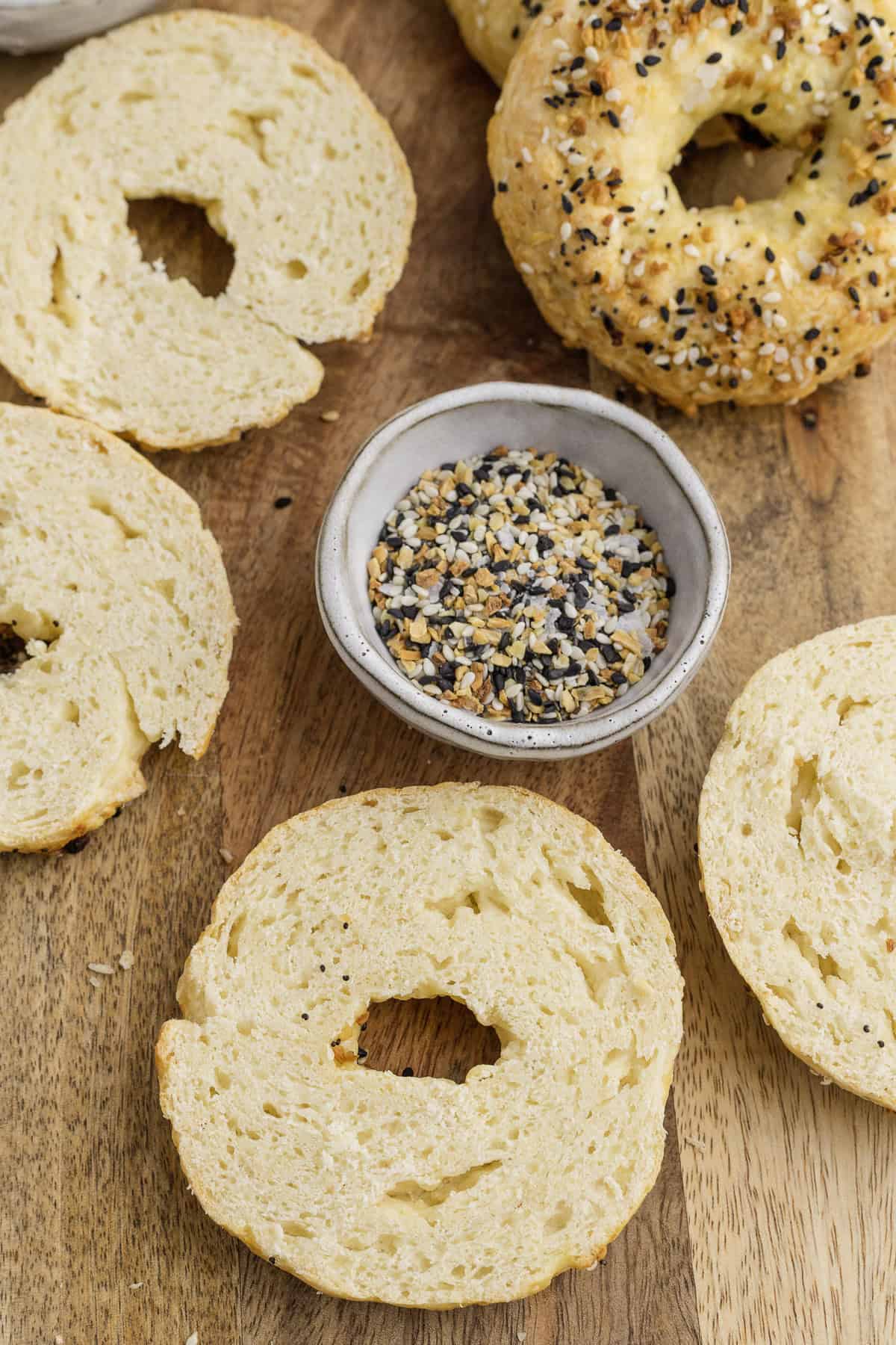 Sliced plain and everything bagels are arranged on a wooden surface, with a small bowl of everything bagel seasoning in the center.
