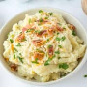 Bowl of mashed potatoes topped with shredded cheese, bacon pieces, and chopped parsley.