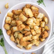 A white bowl filled with golden, seasoned croutons sits on a marble surface, with parsley and a striped towel nearby.