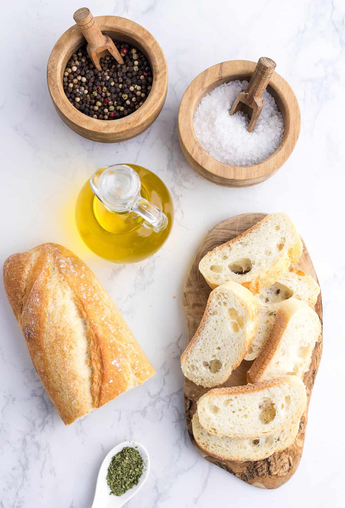 Sliced baguette on a wooden board, whole bread loaf, bowls of mixed peppercorns and salt, a small jug of olive oil, and a spoon of dried herbs on a white surface.