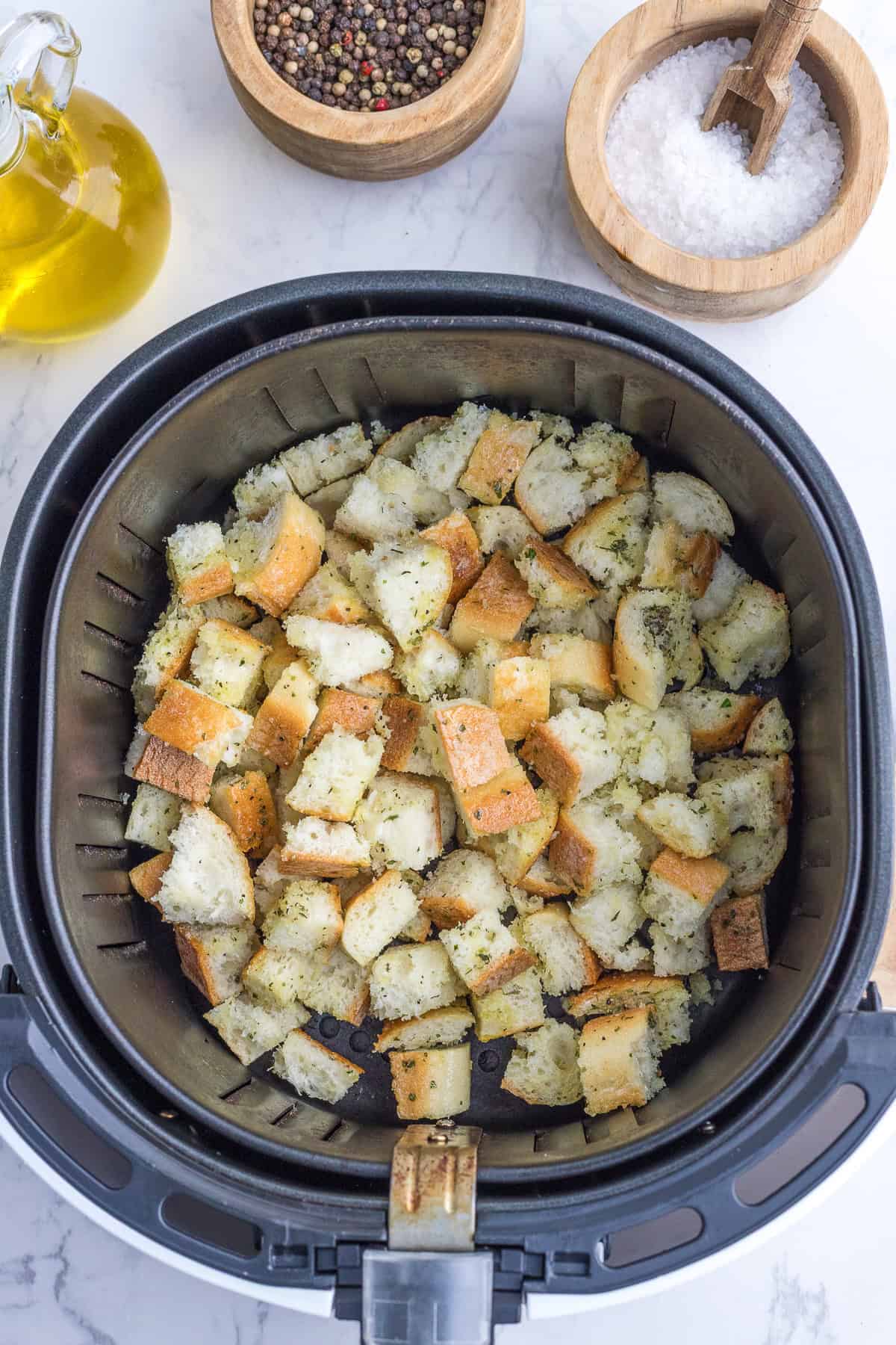 Cubed bread pieces seasoned with herbs in an air fryer basket, with bowls of peppercorns, salt, and a bottle of oil on a marble countertop.
