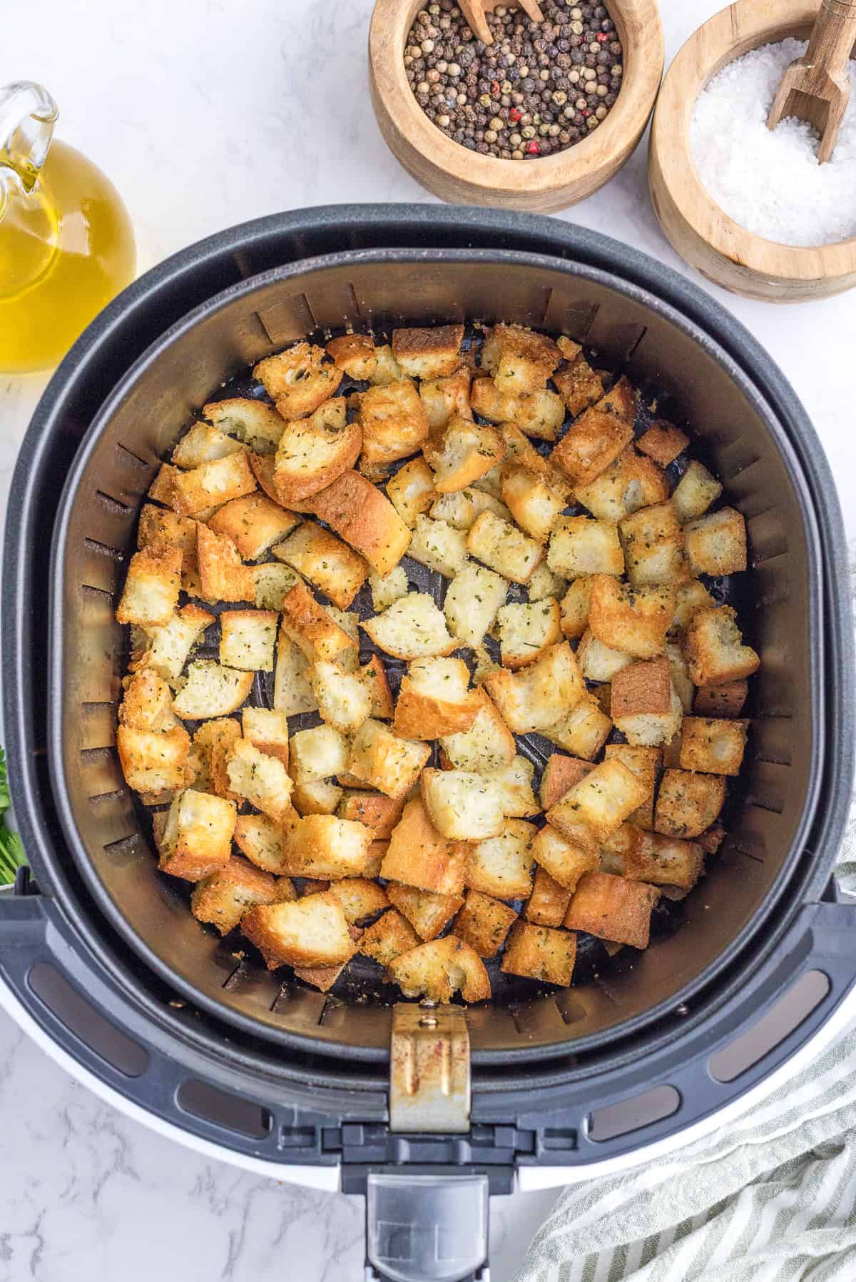 Cubed bread seasoned and cooked in an air fryer basket, with bowls of pepper, salt, and a bottle of olive oil visible nearby.
