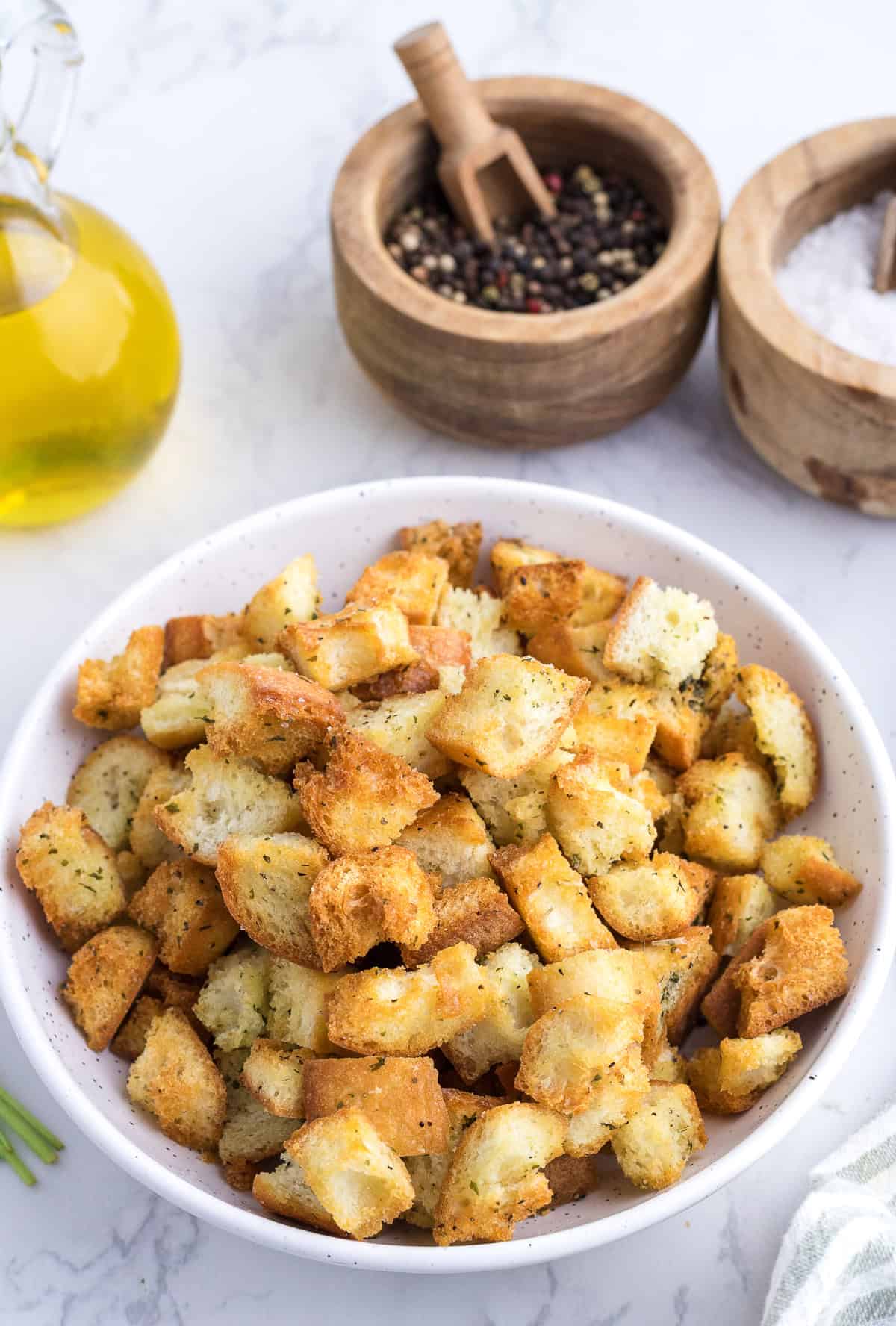 A white bowl filled with seasoned croutons sits on a marble surface near containers of black pepper, salt, and a glass bottle of olive oil.