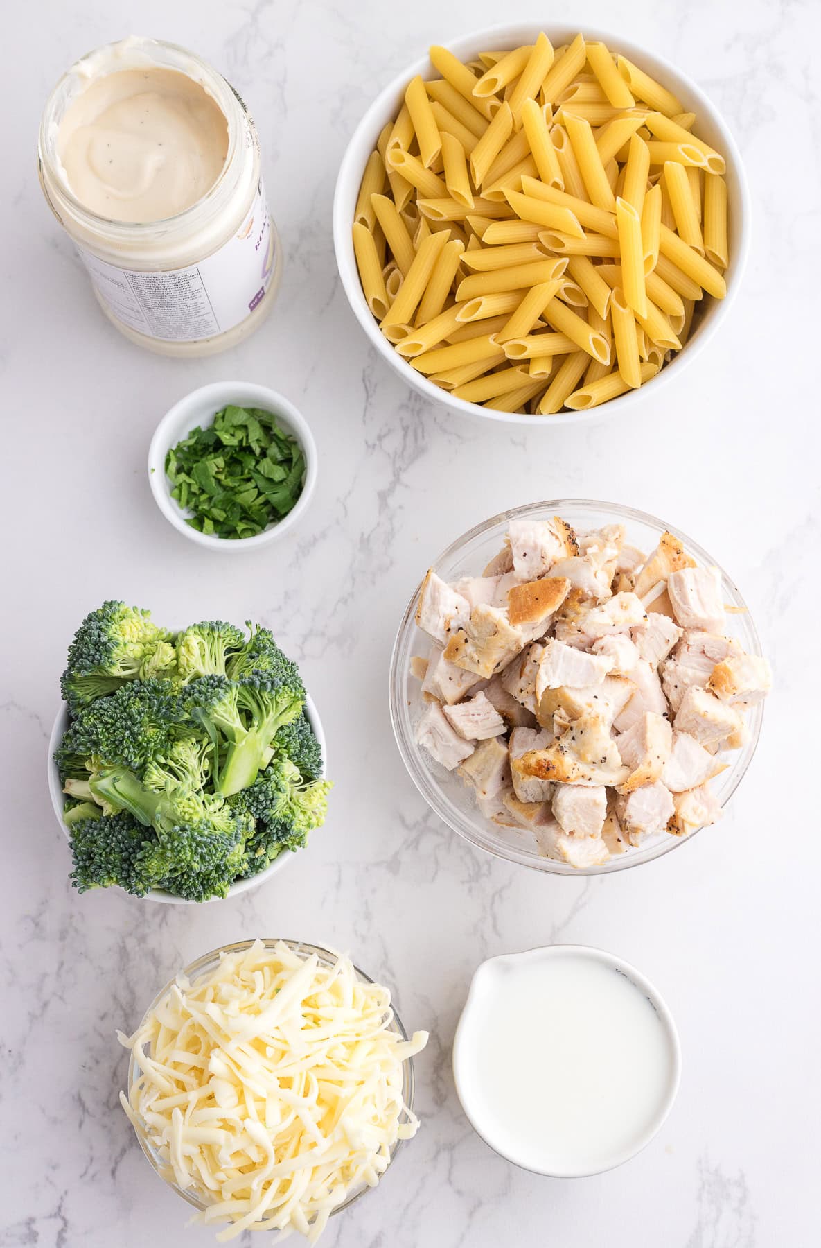 Bowls of uncooked penne pasta, chopped grilled chicken, broccoli florets, shredded cheese, milk, parsley, and a jar of Alfredo sauce arranged on a white surface.