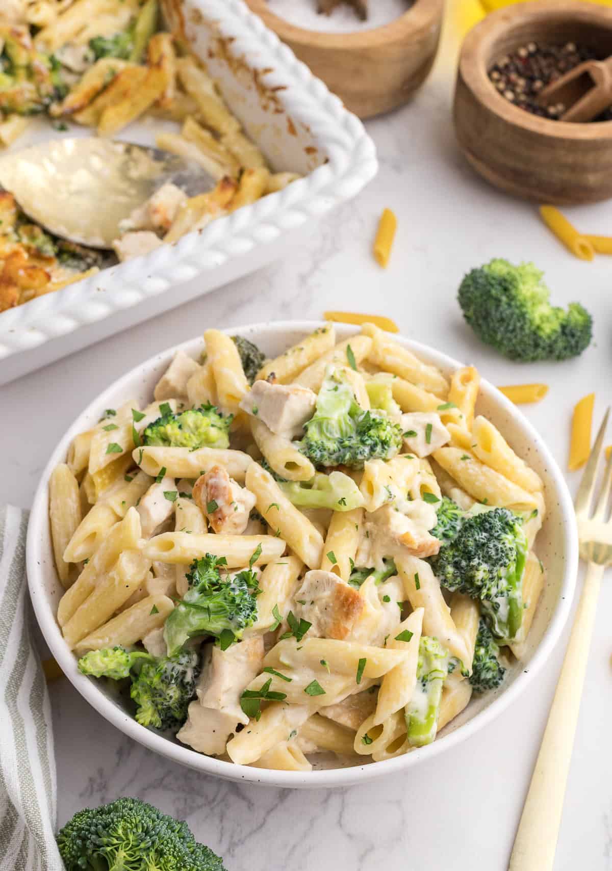 A bowl of penne pasta with broccoli and chicken in a creamy sauce, garnished with chopped parsley. A baking dish and small bowls of spices are in the background.
