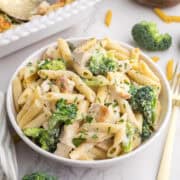 Bol de pâtes penne avec des morceaux de poulet, des brocolis et une sauce crémeuse, agrémentée d'herbes hachées. Une fourchette et des pâtes non cuites se trouvent à proximité.