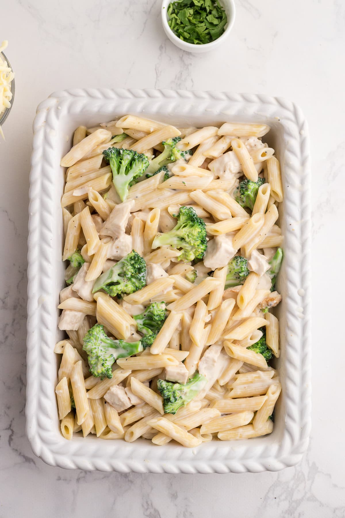 A white ceramic dish filled with penne pasta, broccoli florets, and chunks of chicken in a creamy sauce, with a small bowl of chopped herbs nearby.