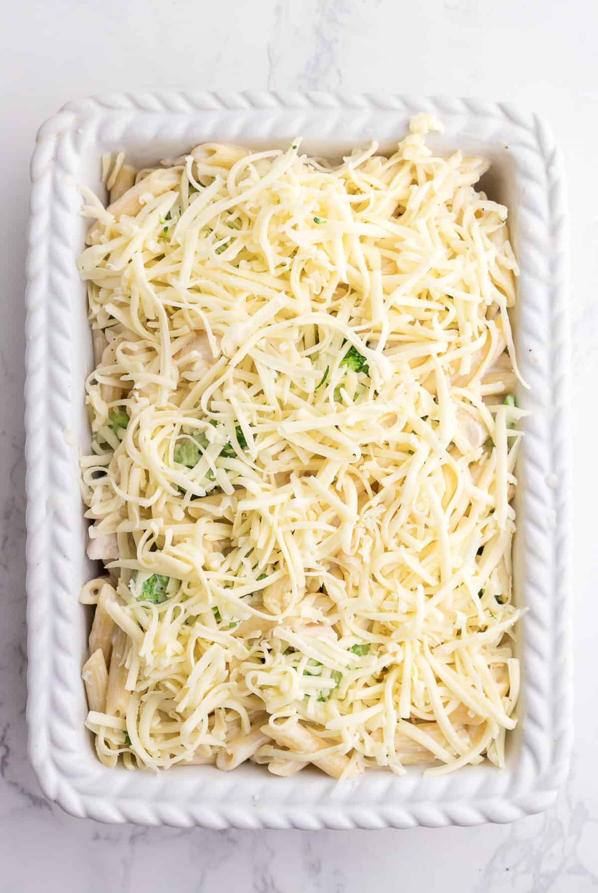Rectangular white baking dish filled with pasta mixed with broccoli and topped with shredded cheese, set on a marble surface.
